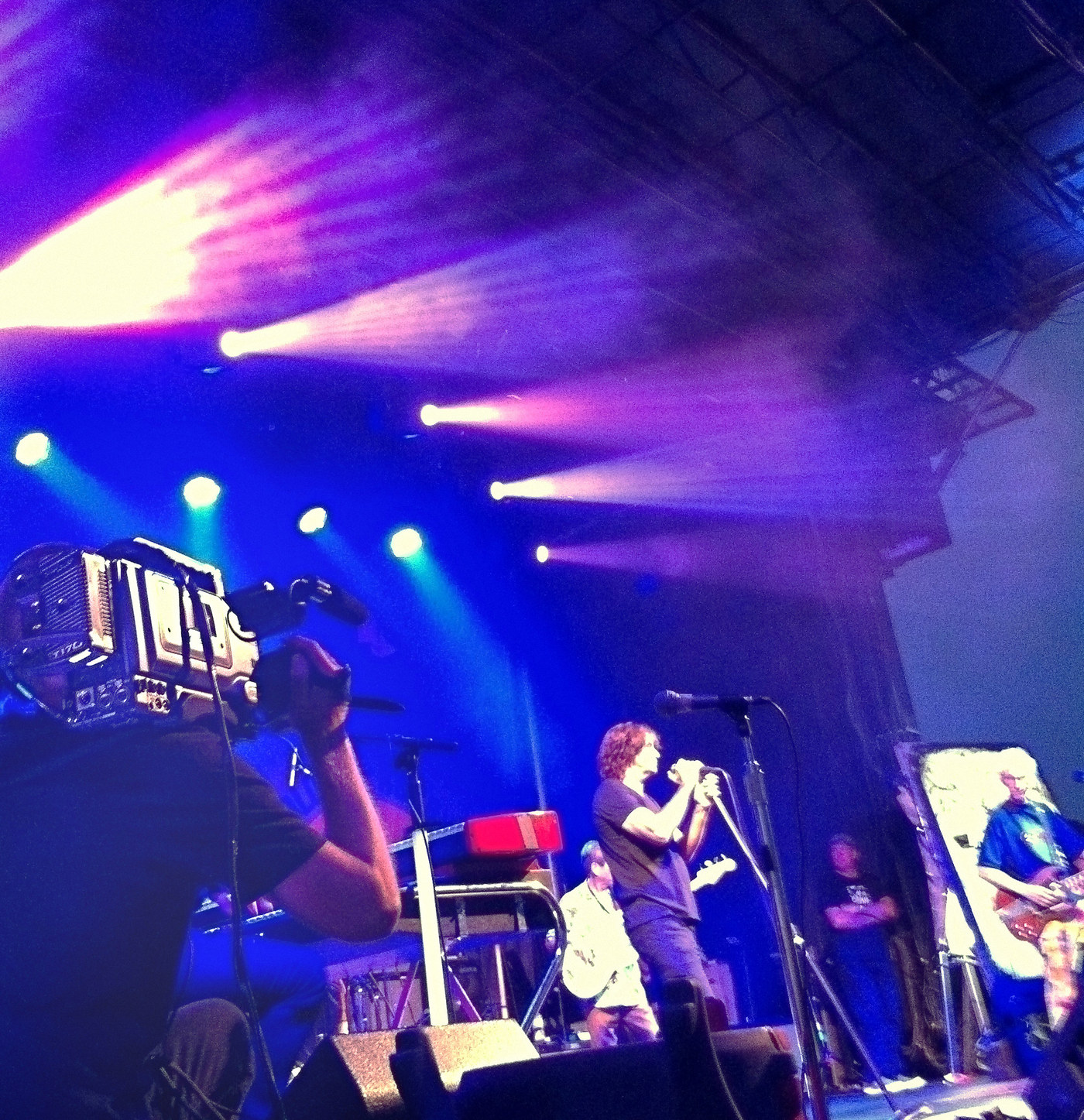 A camera operator working a broadcast camera at the downstage edge of a concert stage, singer at the microphone and band behind under blue and magenta beams.