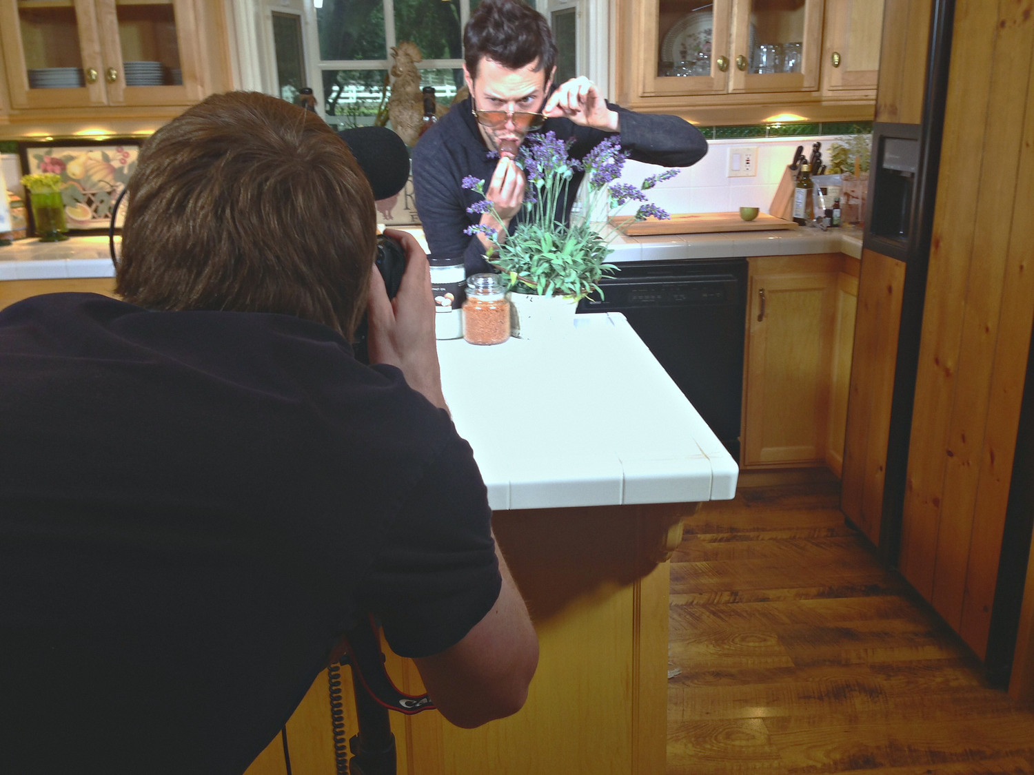 A photographer shooting stills across a kitchen counter dressed with lavender and jars, presenter performing behind it, microphone in frame.
