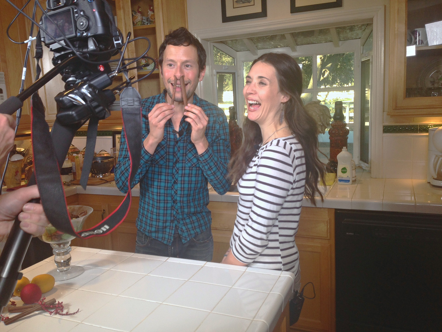 Two presenters laughing on a dressed kitchen set, with a camera on a tripod and a boom microphone in the foreground.