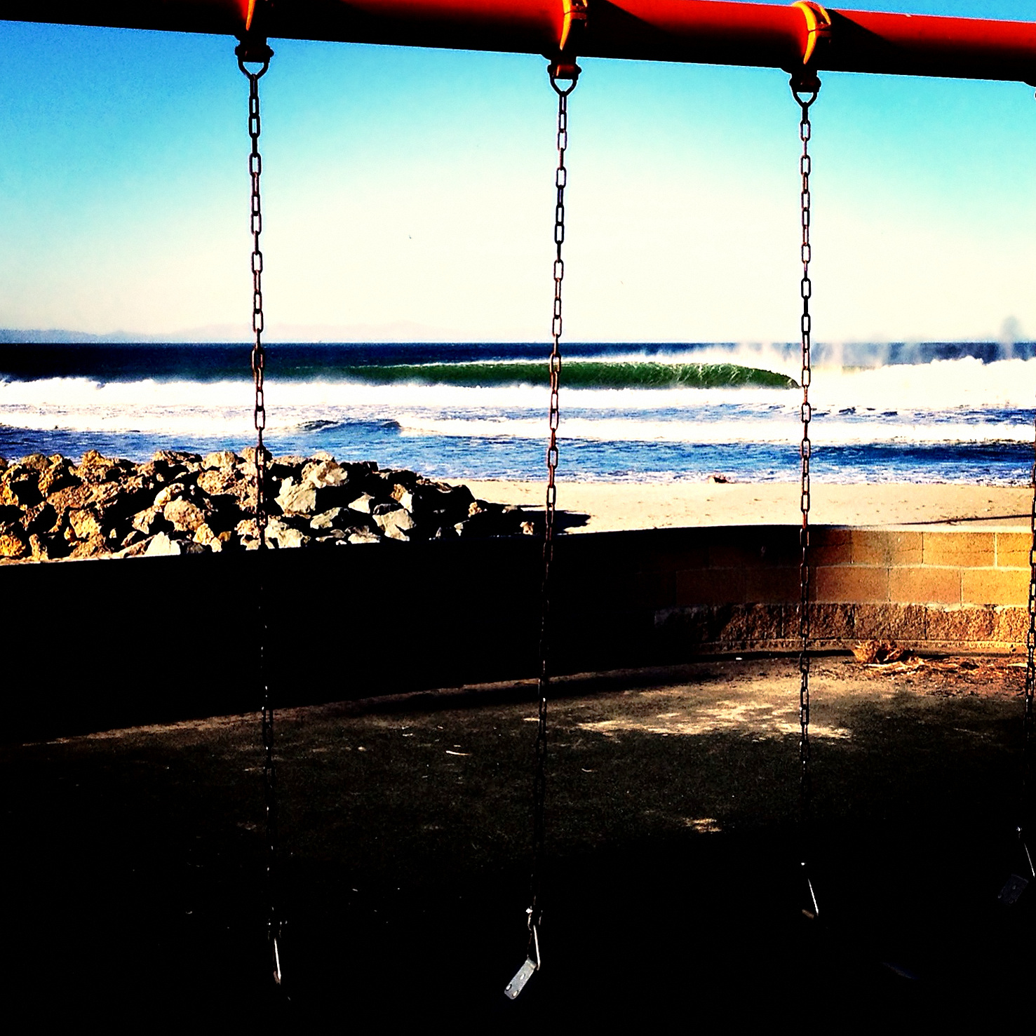 Empty playground swings in silhouette, a clean wave breaking beyond the rock jetty and beach, Ventura, California.