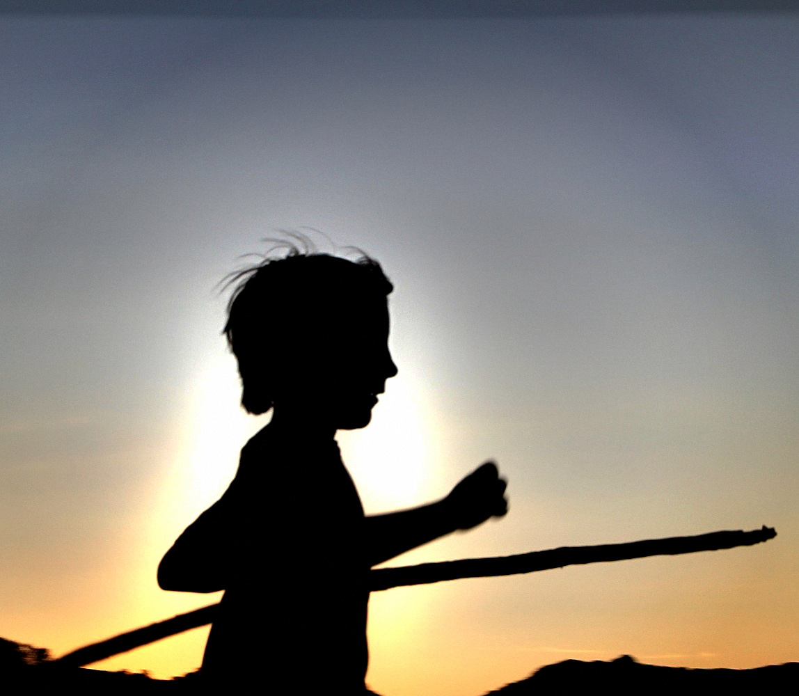 A child in silhouette striding at dusk carrying a long stick, backlit by the last light over a ridgeline, Ventura, California.
